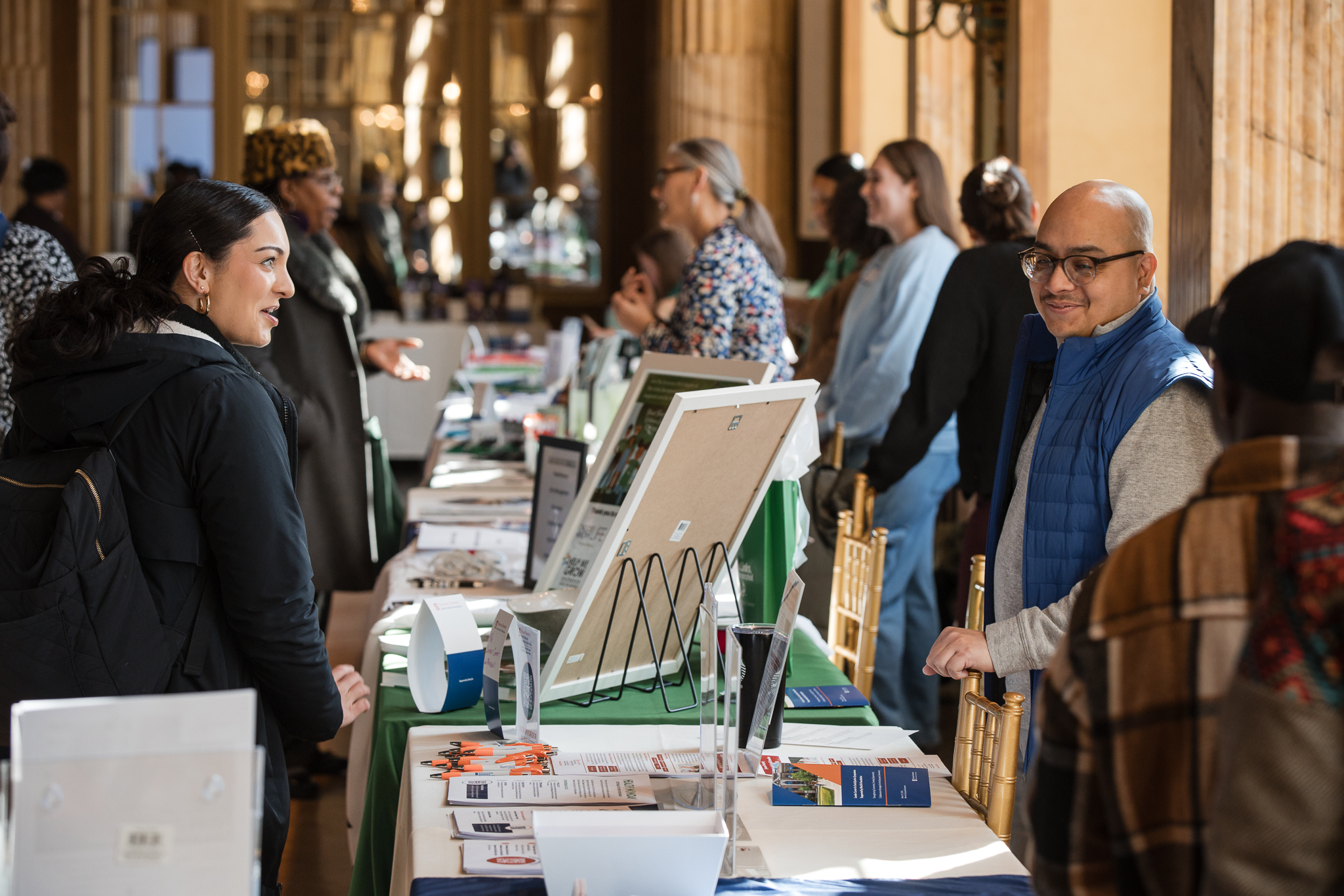 The image shows a busy indoor event with multiple people interacting at various booths. Tables are covered with informational materials, brochures, and display stands. The setting appears to be a conference or fair in a well-lit room with wooden decor and large windows.