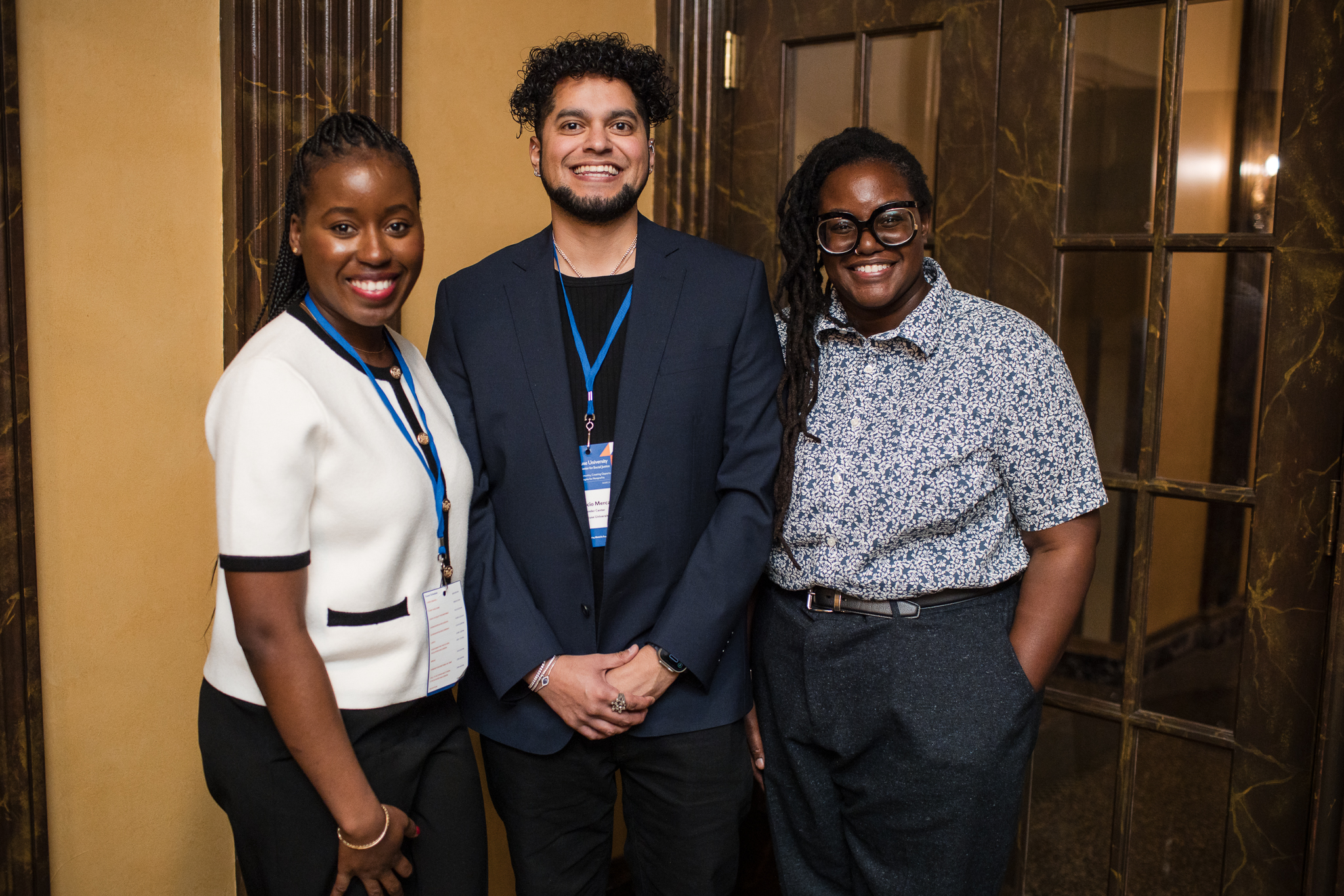 The image shows three individuals standing together in front of a wooden door with glass panels. The person on the left is wearing a white top with black trim and dark pants, the person in the middle is wearing a dark suit with a blue lanyard and badge, and the person on the right is wearing a patterned shirt and dark pants.