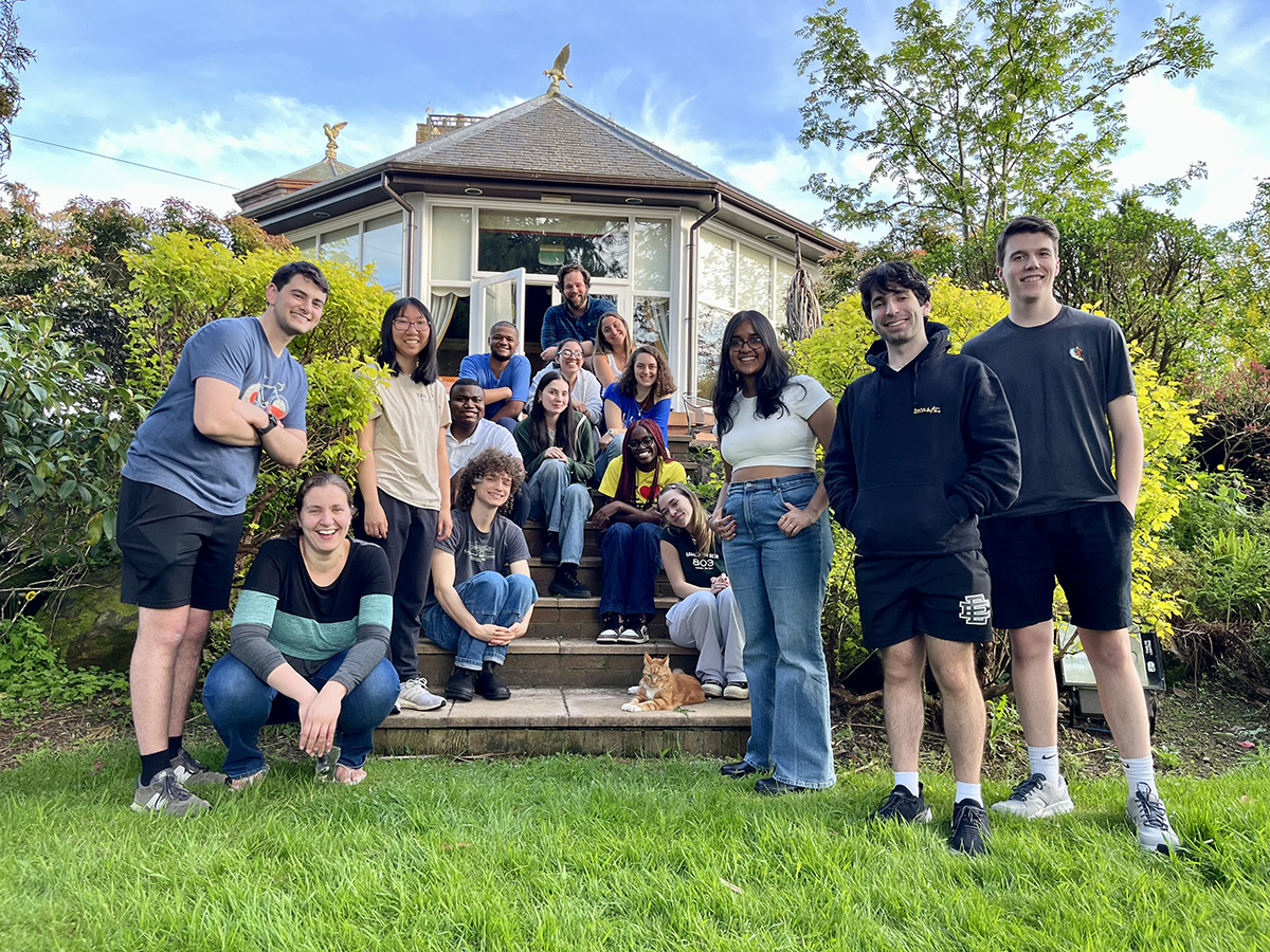 A group of students are gathered outside a house with a conservatory while studying abroad in London. They are posing on and around the steps leading up to the house. The background includes greenery and trees, and there is a cat on the steps.
