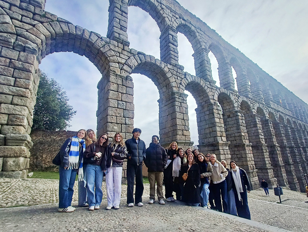 A group of students stand in front of an ancient stone aqueduct with multiple arches while studying abroad in Madrid. The aqueduct is made of large, rectangular stones and extends into the distance. The sky is partly cloudy, and the ground is paved with cobblestones.