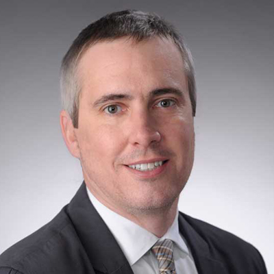 The image shows a person wearing a dark suit jacket, white shirt, and a striped tie against a plain gray background.