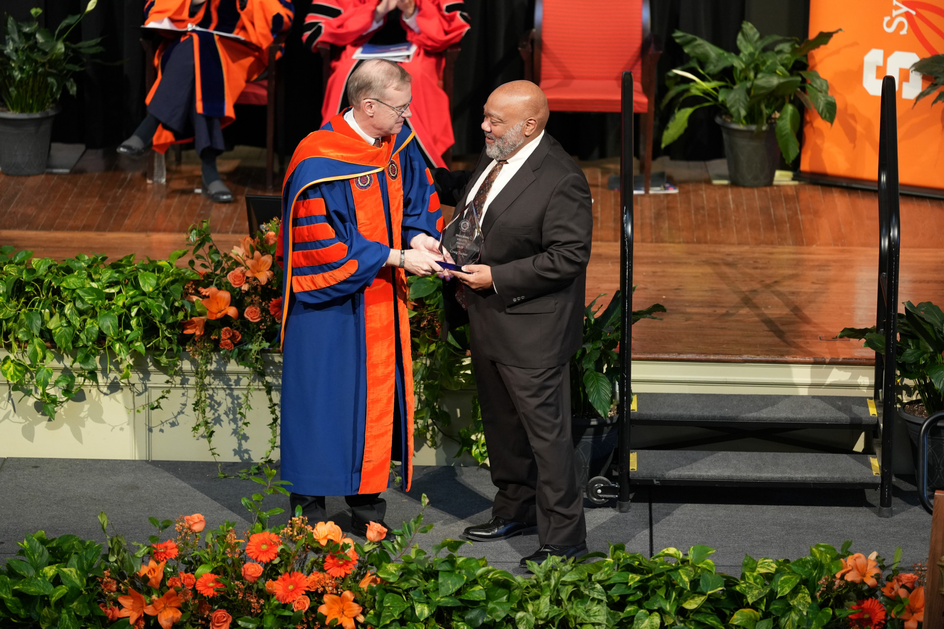 Two individuals are standing on a stage during the One University Awards ceremony. Chancellor Syverud is wearing academic regalia, including a blue and orange gown with a hood, while Craig Tucker is dressed in formal attire, including a dark suit and tie. They are shaking hands, and Tucker is holding an award. The stage is decorated with green plants and orange flowers. In the background, there are other people seated and more decorations visible.