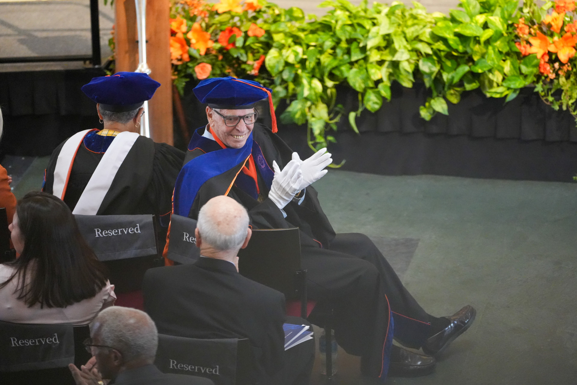 A group of people dressed in academic regalia, including caps and gowns, are seated in a reserved section at an awards ceremony. The individuals are facing away from the camera, except for Professor Samuel Clemence who is applauding and smiling. The background includes a stage with green plants and orange flowers.