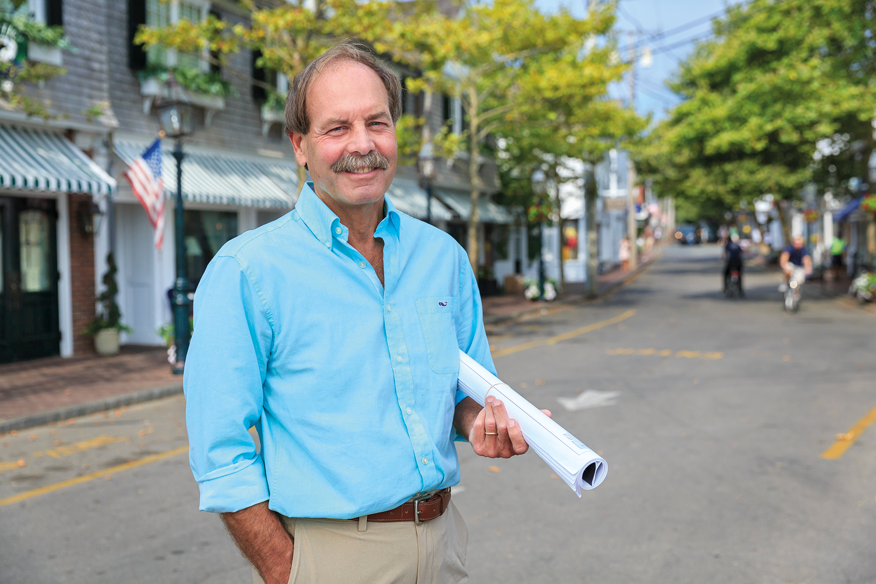Man in a blue shirt, holding rolled-up papers, stands on a street lined with shops and trees.