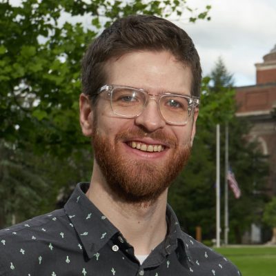 A person standing outdoors. The background includes green trees, an American flag, and part of a brick building. The person is wearing a dark shirt with small cactus patterns and clear-rimmed glasses.