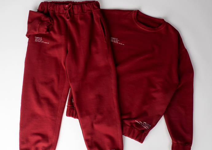 The image shows a matching set of maroon-colored sweatpants and a sweatshirt laid out on a white surface. The sweatpants have an elastic waistband with a drawstring, and the sweatshirt has long sleeves and a round neckline. Both pieces feature small white text printed near the pockets on the sweatpants and near the bottom hem on the sweatshirt.