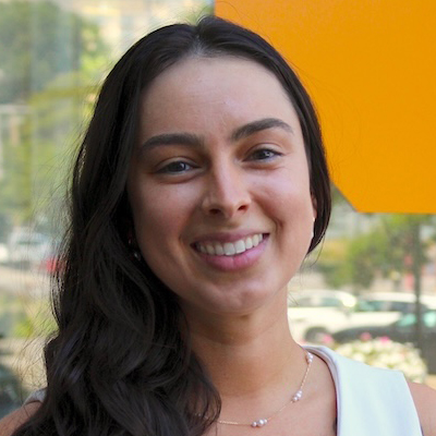 The image shows a person with long, wavy dark hair standing in front of a blurred outdoor background. The person is wearing a light-colored top and a delicate necklace with small beads.