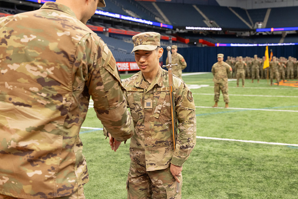 a military ceremony takes place on a football field. Two soldiers in camouflage uniforms are shaking hands in the foreground, with one holding a ceremonial staff. In the background, there are more soldiers standing in formation.