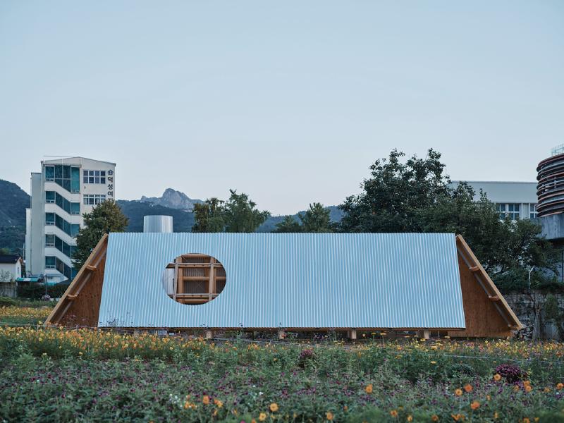 A modern, minimalist building with a corrugated metal roof and wooden supports. The structure features a large circular window on one side. In the foreground, there is a field of wildflowers in various colors. In the background, there are several buildings and trees, with mountains visible in the distance under a clear sky.