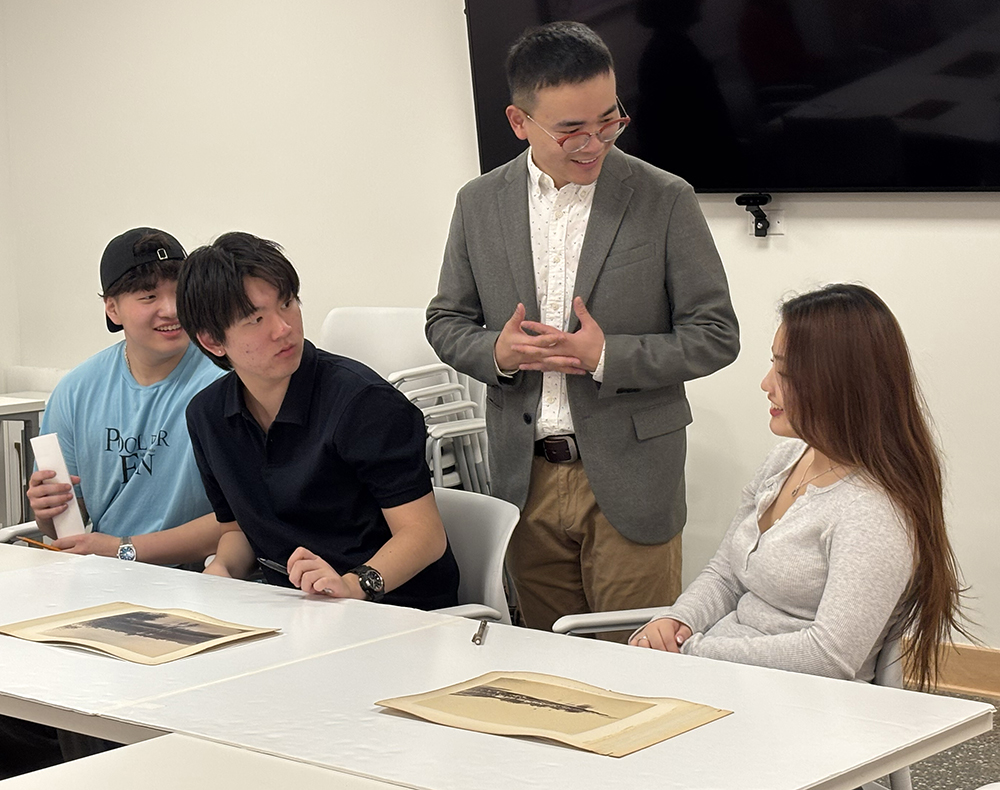 A professor and three students gather in a classroom setting. The students are seated at a table with works of art spread before them, while the professor stands and speaks to them. There is a large television screen mounted on the wall behind them.