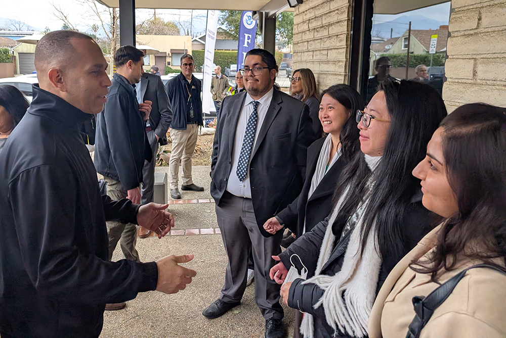 A group of people is standing outside a building, engaged in conversation. The individuals are dressed in business or semi-formal attire, including suits and jackets. One person on the left is gesturing with their hands while speaking to others who are listening attentively. The background shows part of the building's exterior, a covered walkway, and some trees and houses in the distance.
