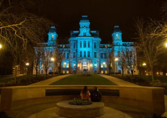 The image shows a large, historic building illuminated with blue lights at night. The building has a central tower and two smaller towers on either side, with numerous windows lit up. In the foreground, there are two people sitting on a circular stone bench facing the building. The surrounding area includes pathways, bare trees, and street lamps providing additional lighting.
