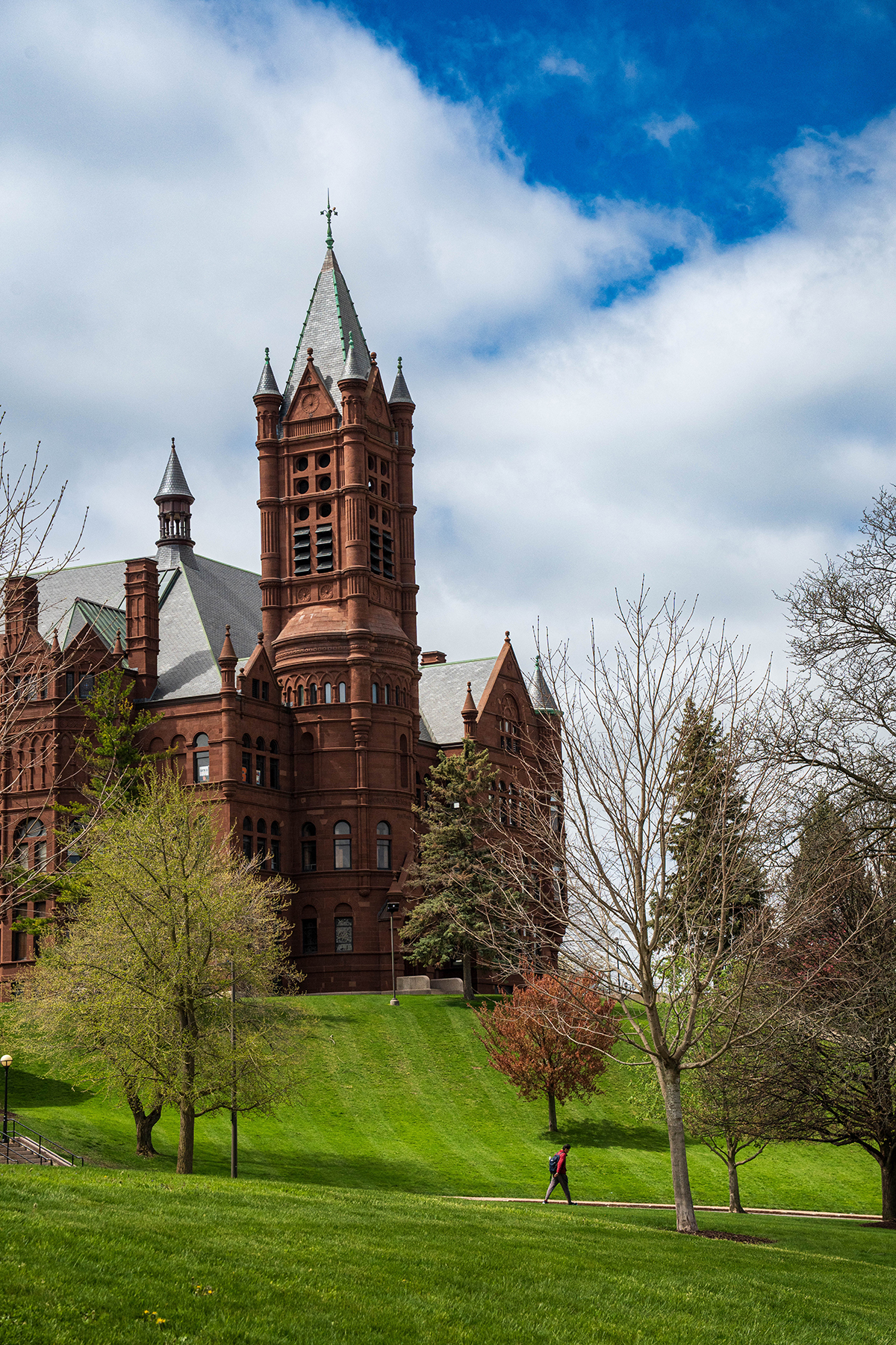 A large historic red-brick building with a tall, pointed tower and several smaller spires, situated on a grassy hill surrounded by trees. A person is walking on a path near the base of the hill under a partly cloudy sky with patches of blue.