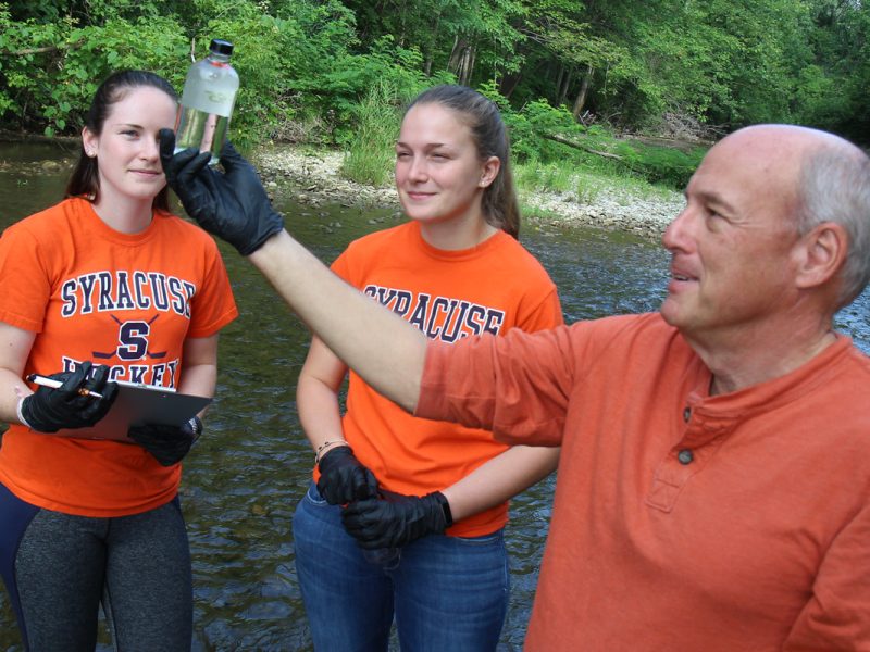 Three people are standing in a shallow river surrounded by greenery. Two of them are wearing orange "Syracuse" t-shirts, and one is holding a clipboard. The third person, wearing an orange long-sleeve shirt, is holding up a small clear bottle filled with water.