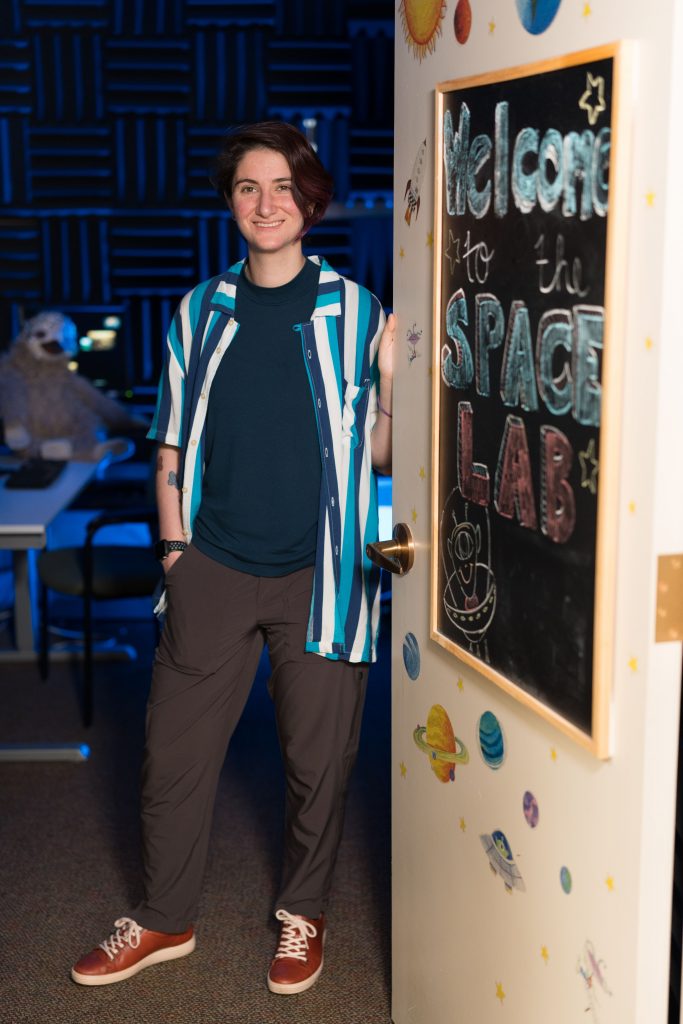 A person is standing in a doorway, leaning against the door frame. The door has a blackboard with colorful chalk writing that says 'Welcome to the SPACE LAB' along with drawings of planets and stars. The room behind the person has acoustic foam panels on the walls and a large st￭ animal sitting at a table.