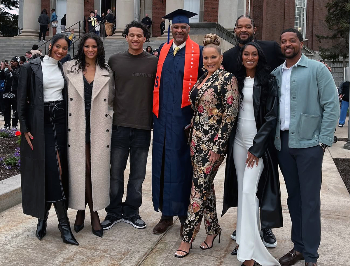 A group of seven people standing together outdoors on Syracuse University's campus following the College of Professional Studies Convocation ceremony. One person in the center is wearing a graduation cap and gown with an orange stole. The other six people are dressed in various styles of clothing, including coats, dresses, and casual wear.