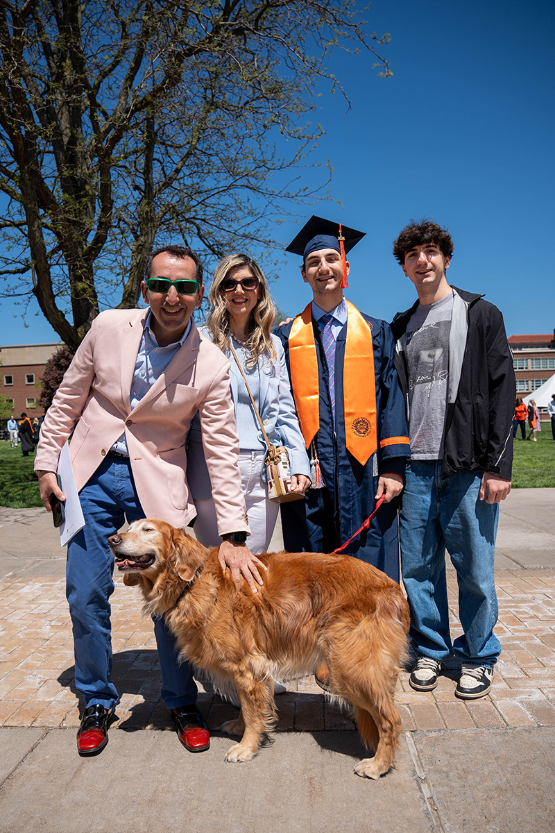 A group of four people standing outdoors during Commencement 2025, with a dog in front of them. One person is wearing a graduation cap and gown with an orange stole. The background includes trees, grass, and buildings.