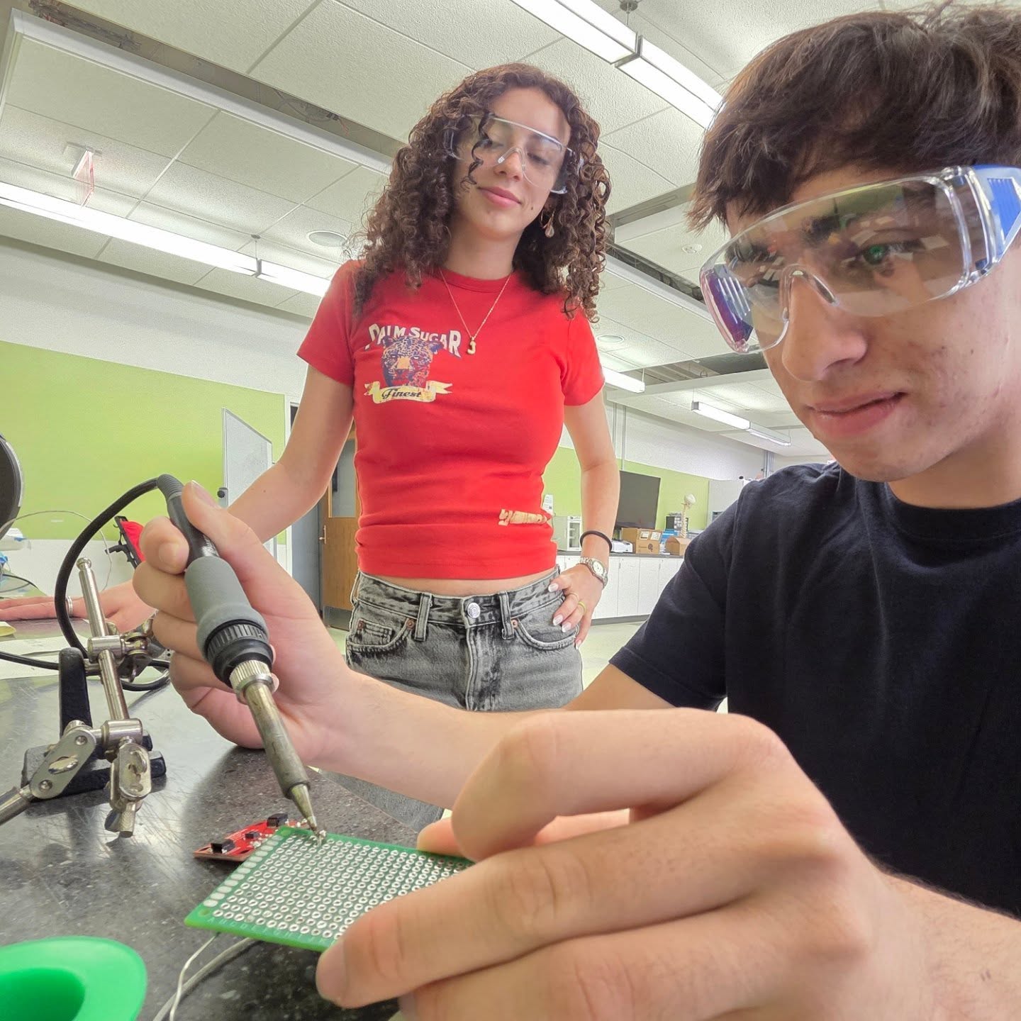 A person using a soldering iron on a green circuit board in a classroom or lab environment, with another individual standing nearby observing. The room has fluorescent lighting, green and white walls, and various pieces of equipment on tables in the background.