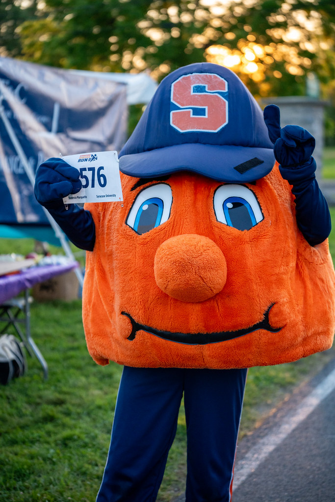 Otto the Orange holds a race bib numbered 576. The background includes trees and an outdoor table.