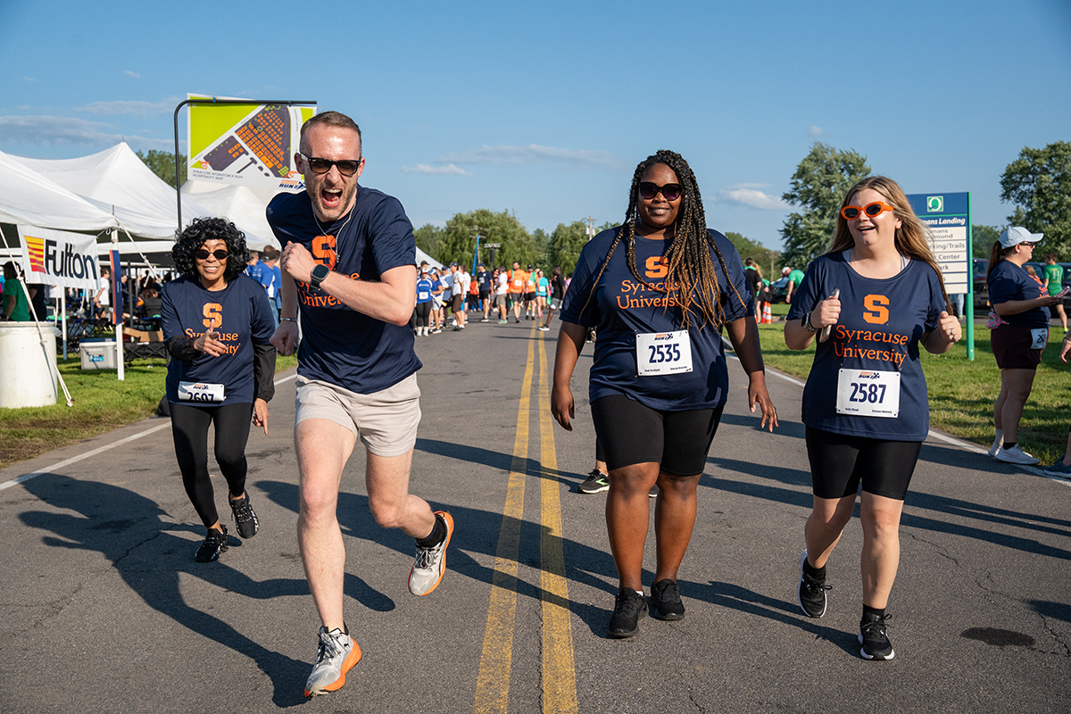Four individuals run on a road during a race event, wearing Syracuse University t-shirts and race bibs numbered 2533, 2587, and 2604. Tents and other participants are visible in the background on a clear, sunny day.