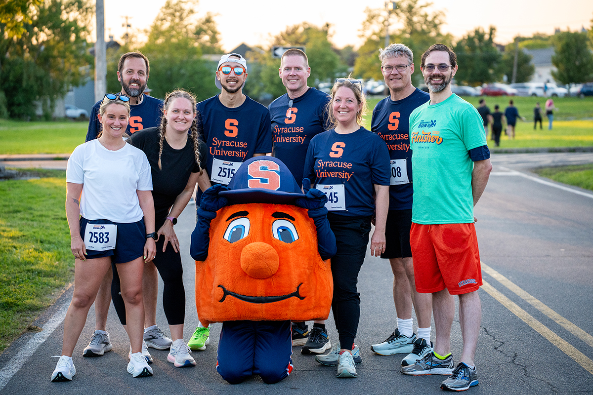 A group of people pose outdoors on a road wearing athletic clothing and race bibs, including numbers 2583, 2515, and 2508. The Syracuse University mascot, Otto the Orange, kneels in front. Several individuals wear shirts with the letter 'S' and 'Syracuse University' text.
