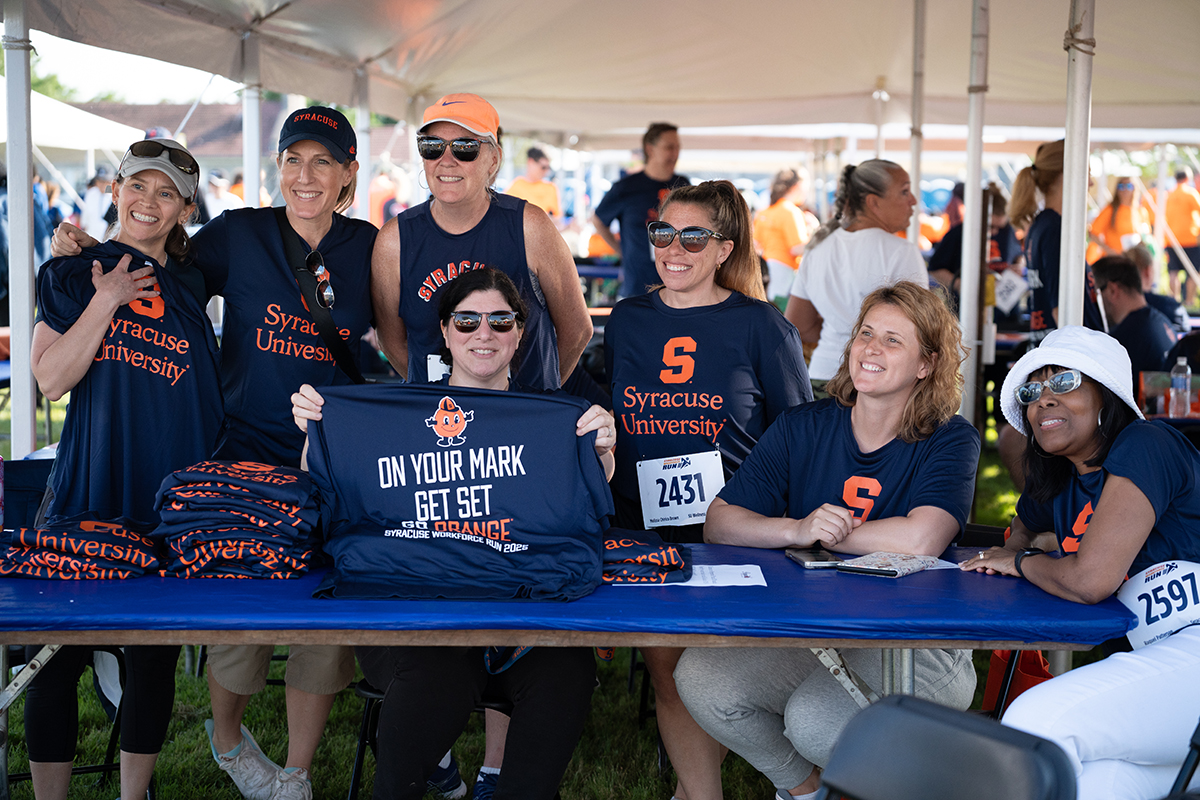 A group of people gather under a tent at the Syracuse WorkForce Run, distributing Syracuse University t-shirts. One person holds up a shirt that reads: 'ON YOUR MARK GET SET GO ORANGE.'