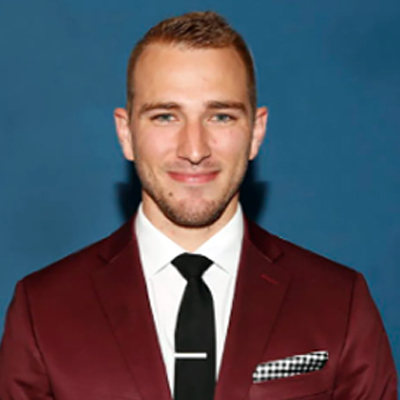 A person is dressed in a burgundy suit jacket, white dress shirt, and black tie. A black and white checkered pocket square is tucked into the jacket. The background is solid blue.