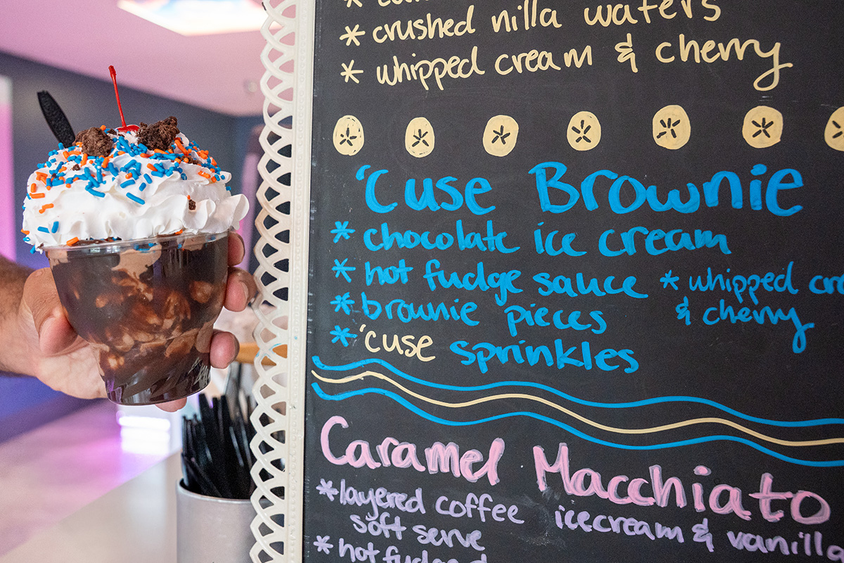 Hand holding a dessert cup with chocolate ice cream, brownie pieces, hot fudge sauce, whipped cream topped with blue and orange sprinkles, an Oreo cookie, and a cherry; beside a chalkboard menu featuring the 'Cuse Brownie and other ice cream items.