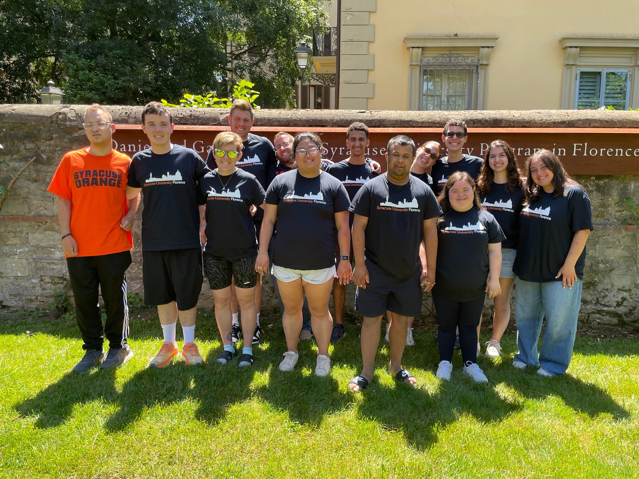 A group of 13 people stands on a grassy area in front of a stone wall with a sign that reads 'Daniel and Gayle D'Aniello Syracuse University Program in Florence.