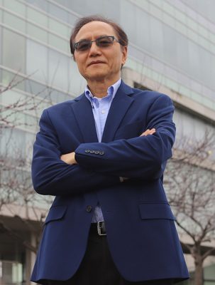 Person in blue blazer and sunglasses standing with crossed arms in front of a modern glass building