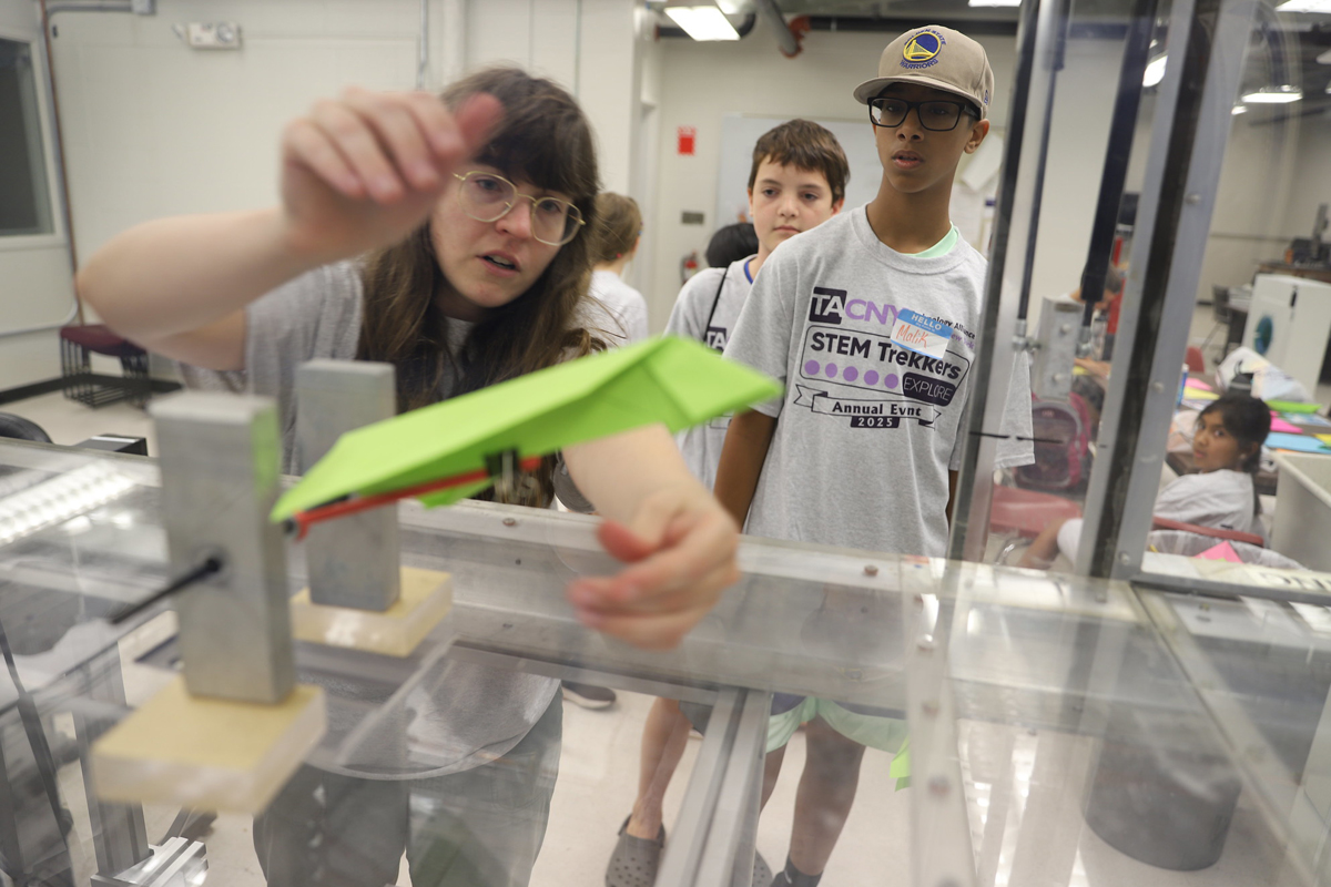 A person with glasses and long brown hair reaches toward a bright green paper airplane or similar object inside what appears to be a clear plastic testing chamber or wind tunnel. They are wearing a gray shirt and is focused on the experiment. Behind her, two other students observe - one wearing a Golden State Warriors cap and a "STEM Trekkers" t-shirt, and another student in the background. The setting appears to be a science laboratory or classroom with white walls, fluorescent lighting, and various equipment visible.
