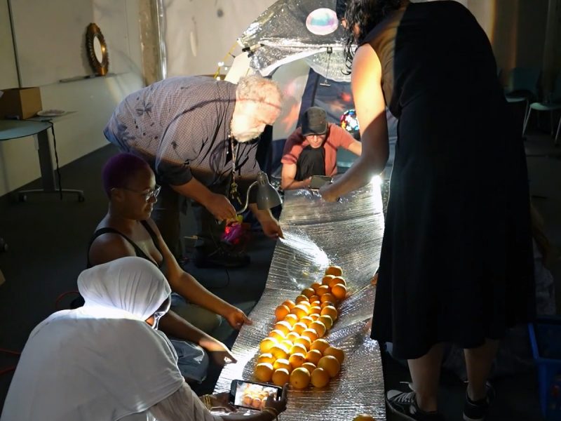 People arranging oranges in a line on a reflective surface indoors, surrounded by chairs, a basket, and a blue crate, with one person capturing the scene on a smartphone.