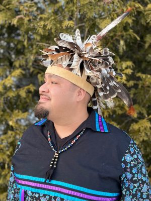 A person wearing a feathered headdress, black shirt with blue and purple patterned sleeves and a beaded necklace stands in front of green foliage.