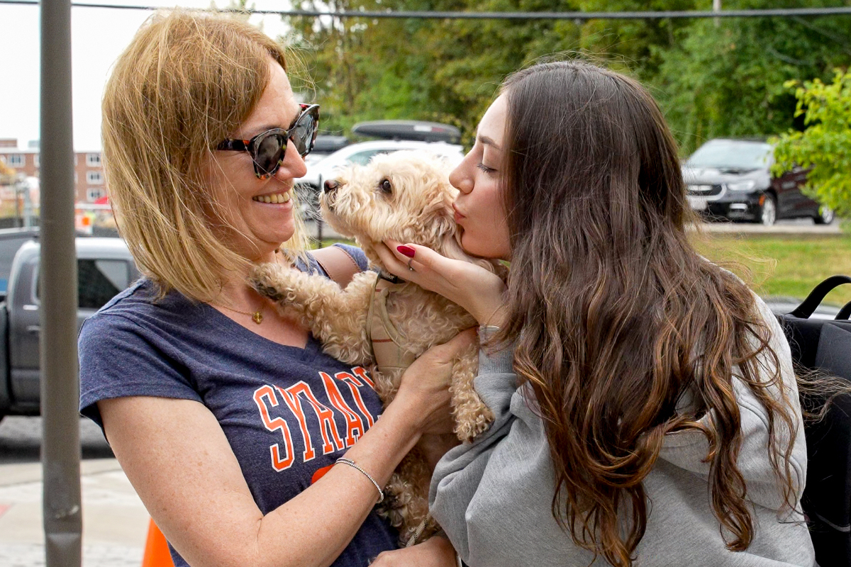 Two people sharing a joyful moment with a small curly-haired dog outdoors. The woman on the left wears sunglasses and a navy "Syracuse" t-shirt, while the woman on the right has long wavy brown hair and is giving the dog a kiss.