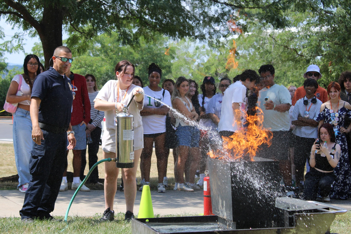 Person demonstrating fire extinguisher use on outdoor flames while a large group observes; safety cones and equipment visible, indicating a fire safety training event.
