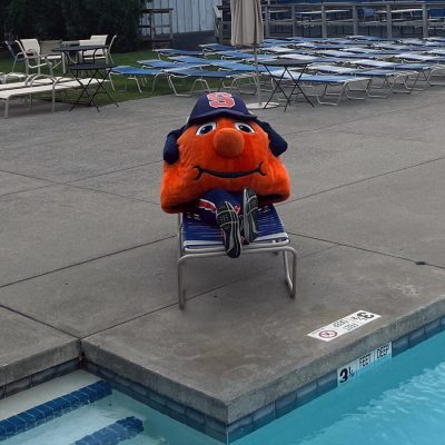 Otto the Orange mascot relaxing on a poolside lounge chair near an outdoor swimming pool