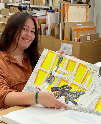 An individual holds a colorful piece of artwork in a storage room at the University's Special Collections Research Center where she is surrounded by boxes and art supplies.