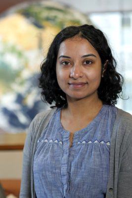 Person with curly hair wearing a blue top and gray cardigan, standing in front of a softly blurred background.