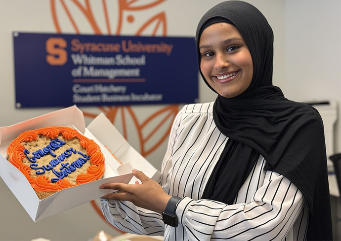 A person in a black headscarf and white striped shirt holds an open box with a cake that has orange frosting and blue writing reading "Congratulations Summer Interns!," in front of a sign for Syracuse University's Whitman School of Management Couri Hatchery Student Business Incubator