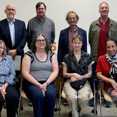 Group photo of eight people posed in two rows, with four seated in front and four standing behind, against a backdrop featuring a bookshelf and partial text.