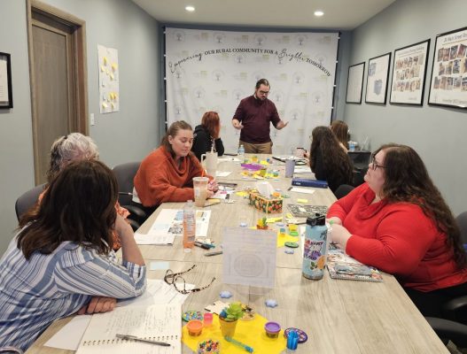 Group of people seated around a conference table with notebooks, drinks, toys, and craft supplies, participating in a workshop led by a presenter. A banner in the background reads 'Empowering Our Rural Community For a Brighter Tomorrow' with 'Family Tree' logos.