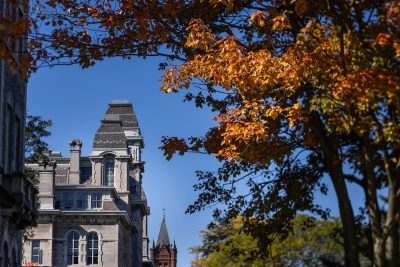 Historic stone building with steep roof and ornate details framed by orange autumn trees under a clear blue sky, with a spired building in the background.