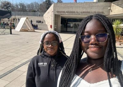 Two individuals stand in front of the Museo de la Memoria y los Derechos Humanos, with steps, people, and a wooden pavilion structure visible in the background.