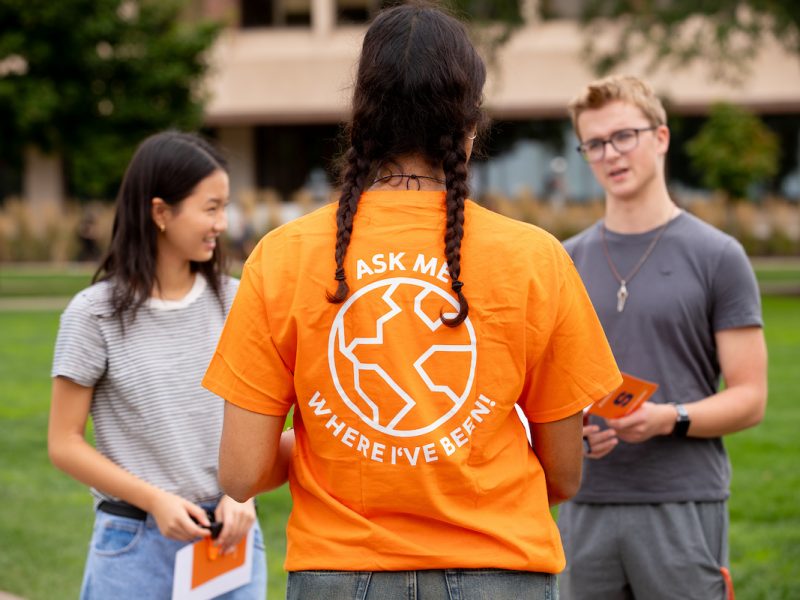 Three people chat with one another outdoors; one wears an orange shirt with 'ASK ME WHERE I'VE BEEN!' and a globe design, facing two others holding pamphlets.