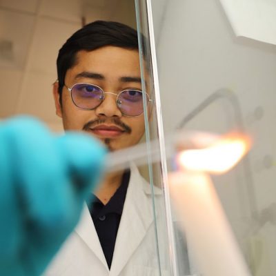 Person in lab coat, glasses and gloves holds a burning candle in a laboratory.