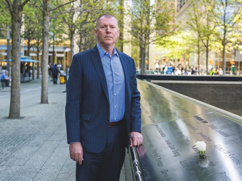 A man in a navy pinstripe suit and blue dress shirt stands solemnly beside the bronze memorial panels at the 9/11 Memorial in New York City, with names of victims engraved in the reflective surface and a white flower placed among them. Trees and urban buildings are visible in the background.