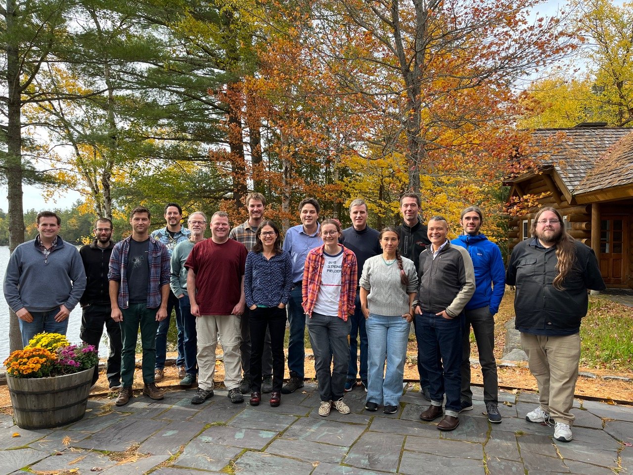 Group photo of approximately 16 people standing on a stone patio in front of a log cabin during autumn. The group includes men and women of various ages wearing casual fall clothing like sweaters, flannel shirts, and jackets. 