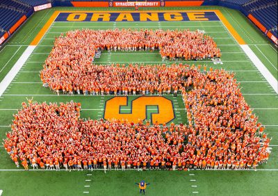 The members of the Class of 2029 in orange clothing form a large "S" on the football field at Syracuse University's JMA Wireless Dome