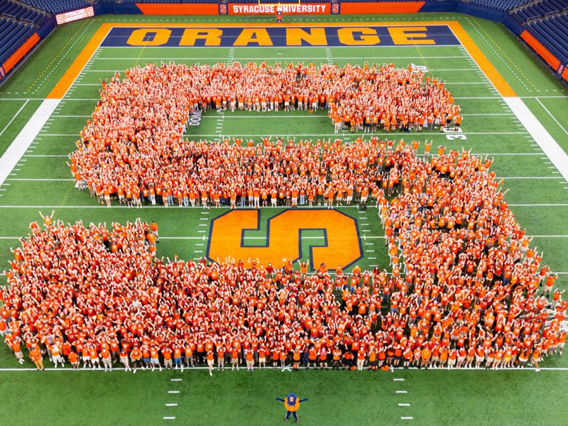 The members of the Class of 2029 in orange clothing form a large "S" on the football field at Syracuse University's JMA Wireless Dome