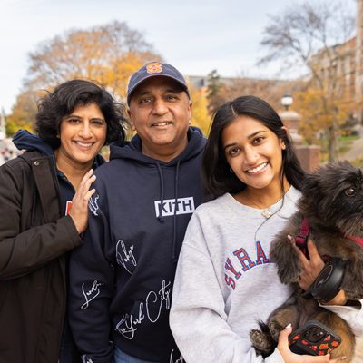 Three people and a small black dog stand on a paved path with autumn trees and buildings in the background.